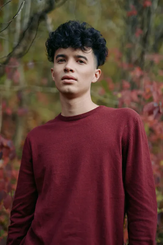 Portrait of a young man in a burgundy sweater standing amid autumn foliage, natural expression and relaxed posture conveying authenticity and credibility in a candid outdoor portrait.