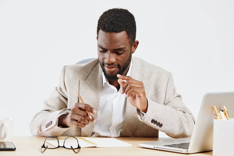 Confident professional man in beige suit with glasses on desk, writing in notebook with laptop and stationery, modern office background, business work lifestyle concept