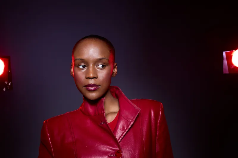 Confident dark skinned African woman with shaved head wearing red leather jacket poses under studio lights on dark background. Concept of fashion, empowerment, beauty, and style.