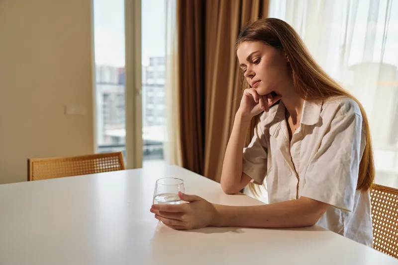 Woman at a table holding a glass, thoughtful portrait in a home interior with warm sunlight. Casual shirt and long hair, calm expression, quiet morning mood and soft natural light.