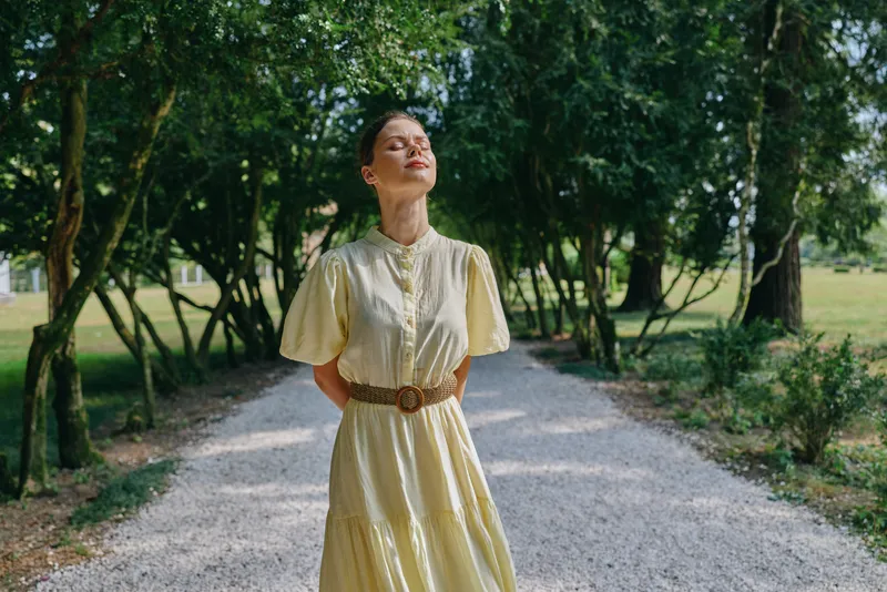 Woman in a yellow dress stands on a gravel path in a park, bathed in sunlight, exuding calm confidence and outdoor style in a natural portrait.