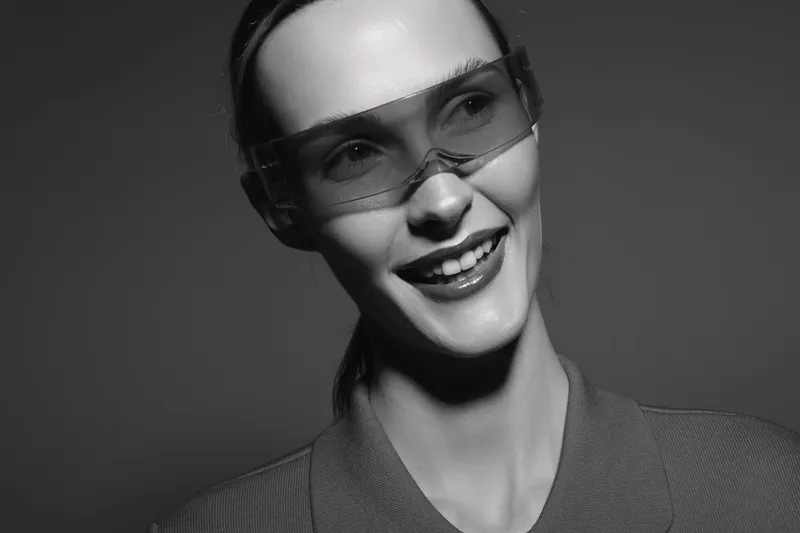 Smiling young white woman with pale skin wearing transparent glasses looks right, showing teeth. Studio portrait in black and white with soft lighting and casual clothing.