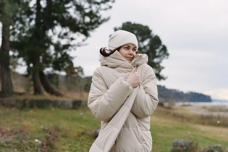 Woman in a beige quilted coat and knit beanie stands in an open outdoor landscape, wrapped in a thick scarf, conveying winter comfort in nature.