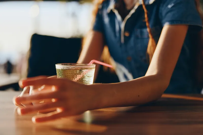 lifestyle woman in modern boho-western denim street style holding a glass with a straw, film color tones capturing a casual and relaxed moment outdoors