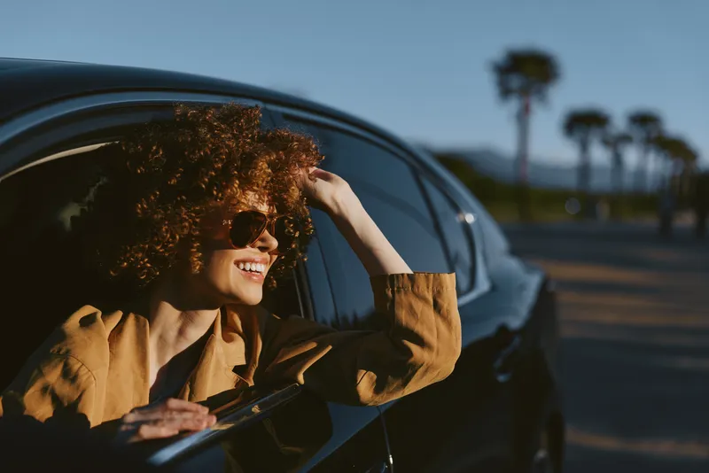 A woman in a camel coat leans out of a sleek car window, smiling behind round sunglasses on a sunny coastal road. A lifestyle moment with retro vintage color tones.