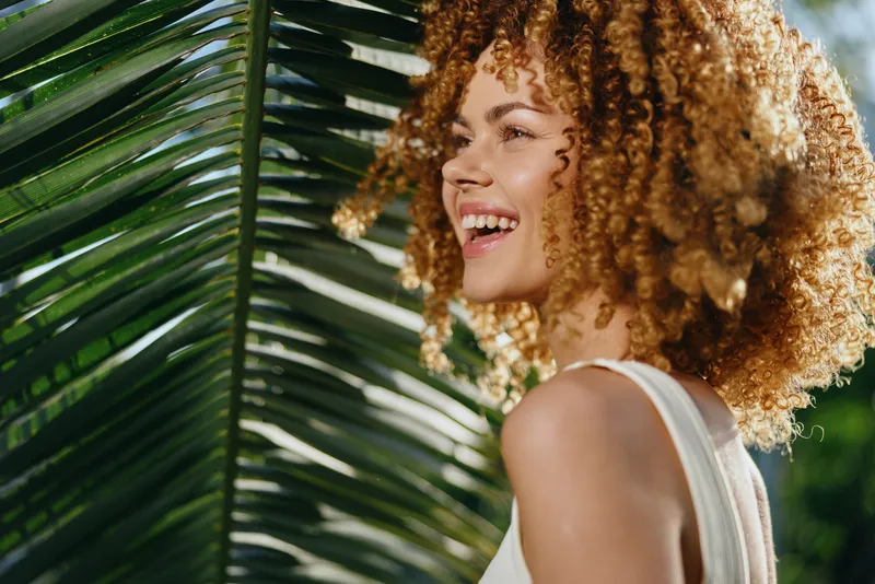 Smiling woman with curly hair enjoying sunny outdoor nature, tropical leaves background, radiant skin and joyful mood in warm daylight, summer lifestyle portrait.