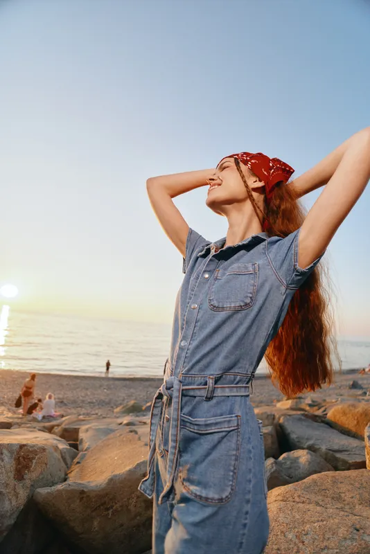 Lifestyle woman in modern boho-western denim street style enjoying sunset by the sea, wearing a red bandana with film color tones and relaxed casual vibe.