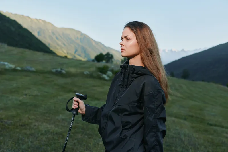 A focused hiker stands in a green valley with distant mountains, gripping a trekking pole. She wears a dark jacket and gazes ahead, conveying adventure and resilience.