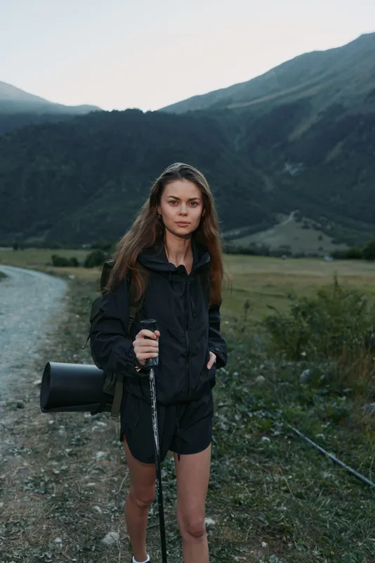 An athletic woman stands alone in a rugged mountain valley, holding a trekking pole and a compact bag, dressed in dark outerwear, gazing toward the distant hills with calm confidence.