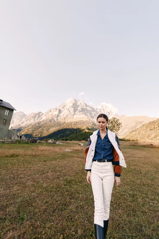 Woman walking in meadow by mountains in countryside wearing denim shirt, layered jacket and boots. Outdoor portrait and travel fashion with serene alpine landscape and warm sunlight.