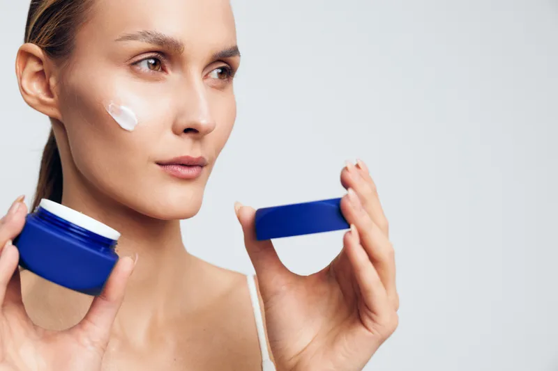Young woman holding blue jar with cream applies moisturizer on face looking away with neutral expression isolated on gray background skincare beauty concept.