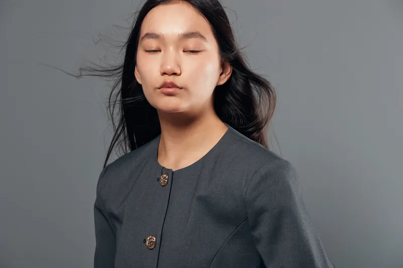 Portrait of young woman with long dark hair and closed eyes wearing a grey jacket on grey background. Calm expression and natural look with minimal makeup and simple hairstyle in studio.