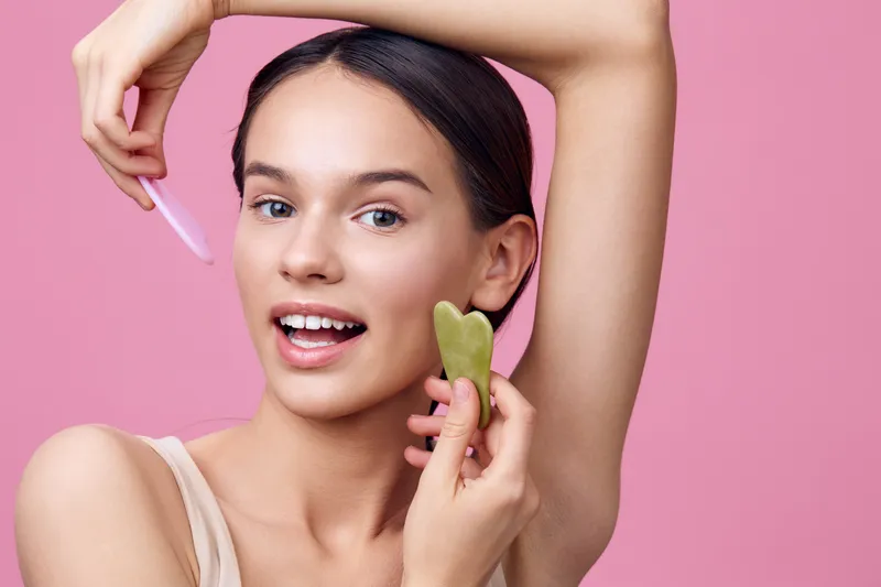 Young woman demonstrating facial massage techniques with gua sha tools on a pink background, promoting healthy skin and self care