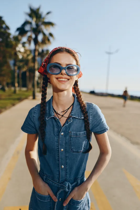Lifestyle woman in modern boho-western denim street style with braided hair and oversized sunglasses, smiling naturally outdoors under bright film color tones.