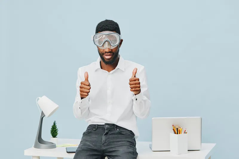 Smiling man with safety goggles in modern office setting, casual white shirt, relaxed posture, thumbs up gesture, work desk with lamp and laptop, positive work environment, professional concept.