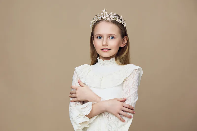 Young girl in a delicate white dress with a crown, exuding elegance and innocence, posed against a soft beige background, perfect for family photography or children s fashion