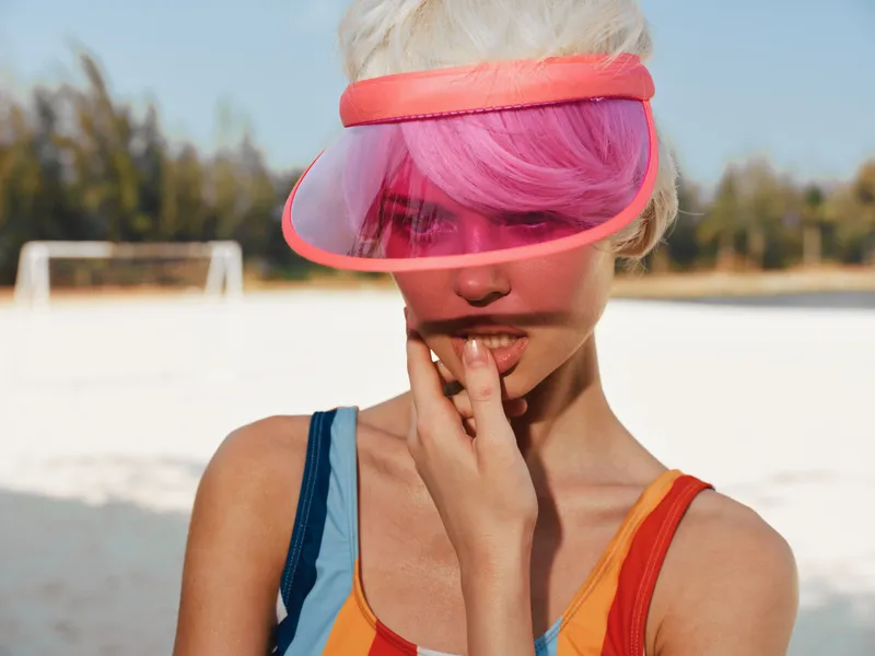 Close up of a stylish young woman with pink hair and a bright pink visor, posing outdoors on a sunny day. She wears a colorful sleeveless top and gazes thoughtfully.