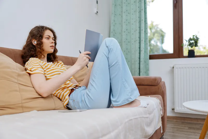 Casual woman relaxing at home on a couch, reading a notebook with a calm expression, wearing a striped shirt and jeans, bright living room atmosphere, lifestyle portrait
