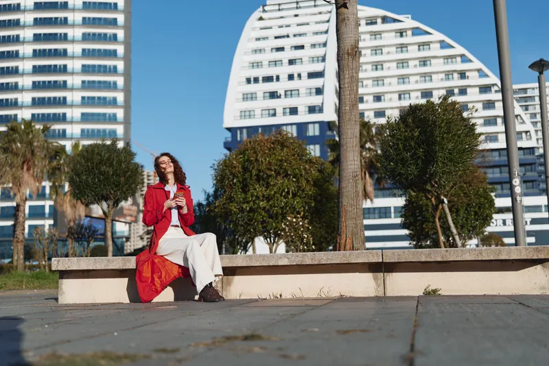 Cheerful woman outdoors in a red coat and white dress sits on a bench in a sunny urban park, modern buildings in background, relaxed pose, bright colors.
