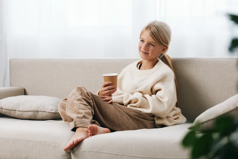 A candid, authentic scene showing play and school lifestyle. A relaxed child sits on a sofa, wearing a cozy sweater, holding a warm cup, expressing genuine mood and everyday comfort.
