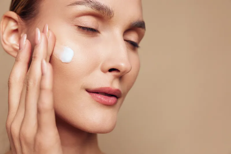 Closeup of a woman applying moisturizer to her cheek, calm expression, smooth skin, skincare routine moment, soft studio lighting, beige background, healthy glow, refreshed and focused