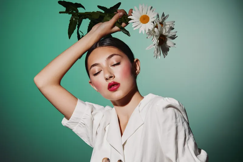 Young woman with bright red lips and flowers on head poses with closed eyes, wearing a stylish white outfit on green background. Fashion beauty concept with creative flower headpiece.