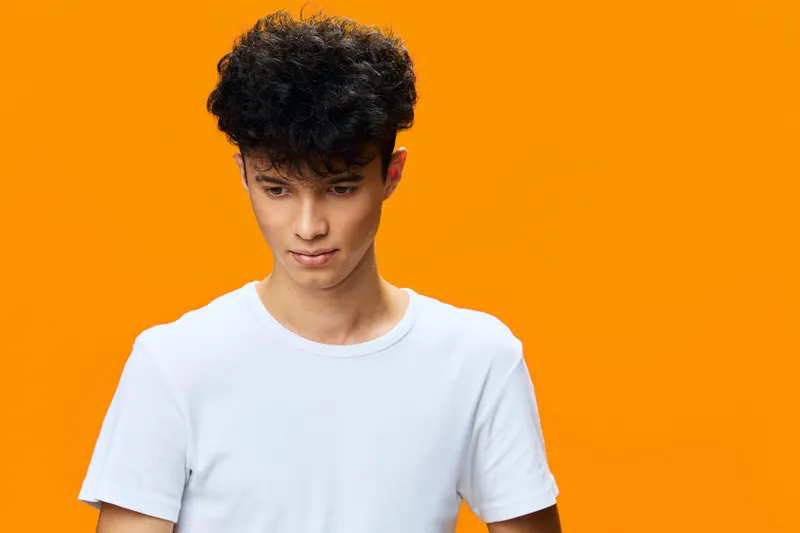 young man with curly hair wearing white t-shirt looks down thoughtfully against vibrant orange background, casual style portrait with bright colors and soft lighting