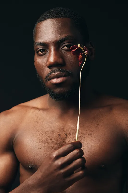 African man with dark skin holding dried flower near face in creative conceptual visual style. Portrait of shirtless black male model with artistic expression on dark background in studio lighting.