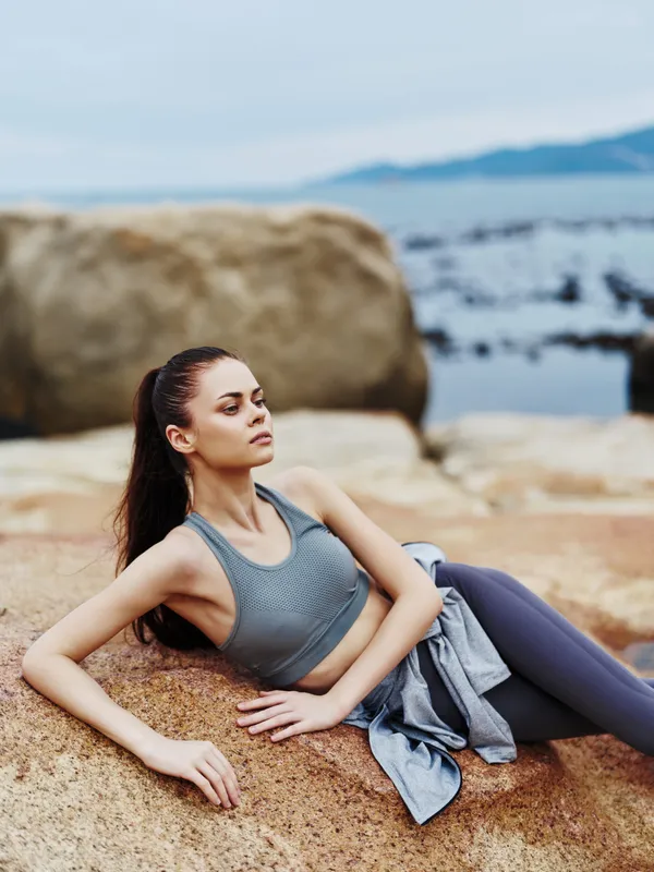 Young woman in athletic wear relaxing on a rocky beach, embodying wellness and peace, with serene ocean background, ideal for healthy lifestyle concepts