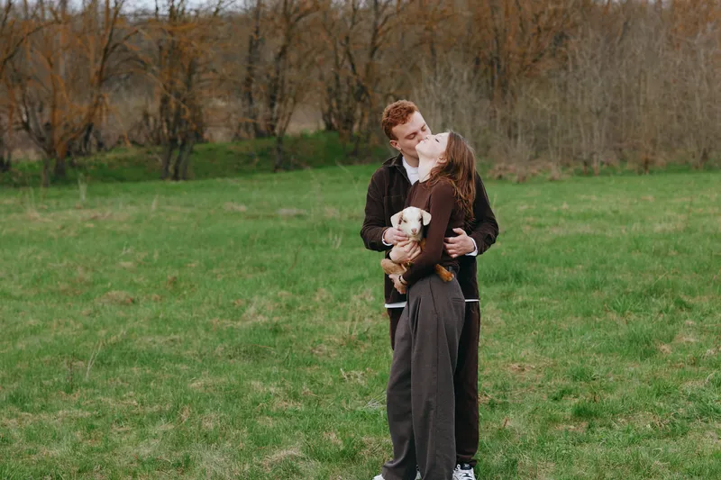 A couple in casual clothing share a tender moment outdoors, holding a small goat kid in a green field. A bright rural scene captures lifestyle, affection, and animal companionship.