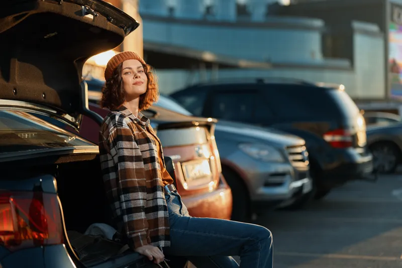 Woman sitting by car trunk in urban parking at golden hour, candid lifestyle portrait with authenticity, golden hour glow, mindful living vibe and emotional storytelling moment.
