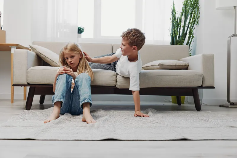 A young boy comforts a sad girl sitting on the floor in a bright living room with a beige sofa and plants. Concept of sibling support, empathy, and emotional connection.