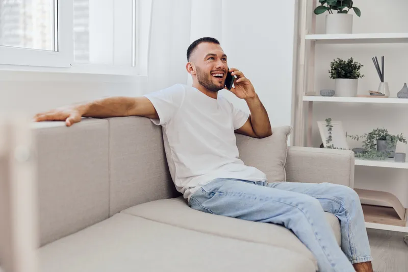 Smiling man engaged in a heartfelt phone call, reflecting emotional vulnerability and modern therapy culture in a cozy home environment.