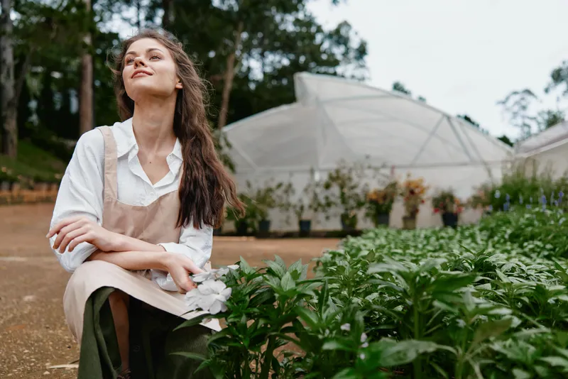 Young woman in an apron enjoying nature in a greenhouse, surrounded by vibrant plants and flowers, showcasing the beauty of gardening and sustainable living