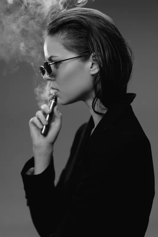 Black and white portrait of a woman vaping in dark clothing and sunglasses with smoke around her, stylish and modern mood.