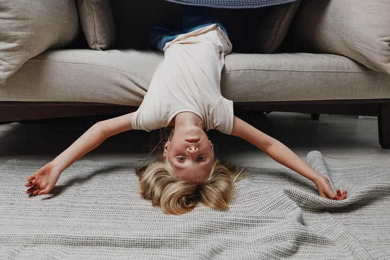 Young boy lying upside down on couch with arms spread on cozy blanket, playful and relaxed mood indoors at home.