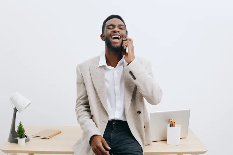 Confident young smiling man talking on mobile phone in modern office, casual business attire, cheerful expression, light background, workspace with laptop and greenery, professional lifestyle concept