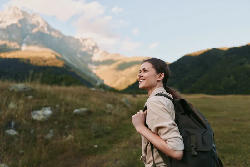 An outdoor woman with a backpack stands in a mountain valley, smiling into the distance. A lifestyle portrait capturing hiking, travel, and nature curiosity.