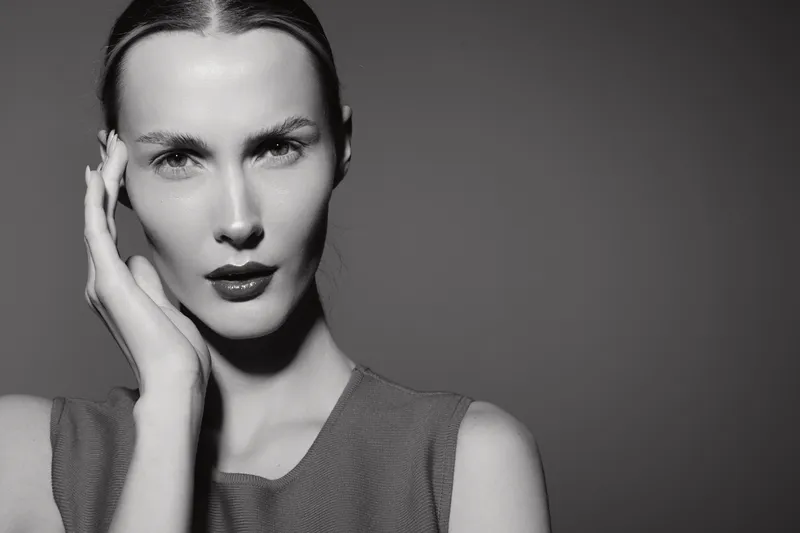 Portrait of a white woman with light skin and serious expression touching her face, studio black and white photo.