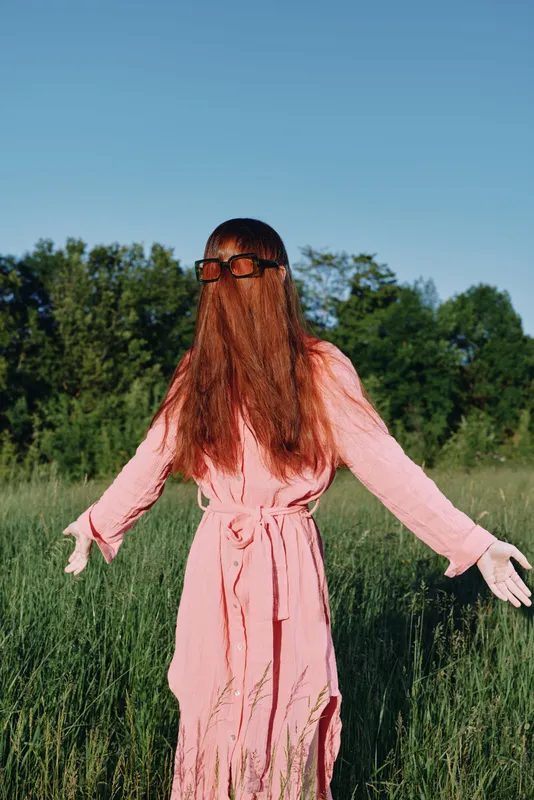 A woman in a pink robe stands in a lush green field, arms outstretched, expressing freedom and joy, with long brown hair flowing in the breeze