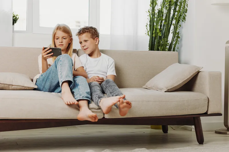 Two children sitting barefoot on beige sofa watching smartphone together in bright living room with plants and cushions. Concept for family, technology, and childhood.