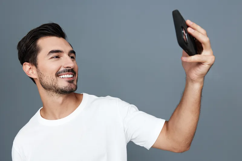 Smiling young man wearing white t-shirt happily taking a selfie with smartphone while standing against gray background. Modern technology lifestyle concept.