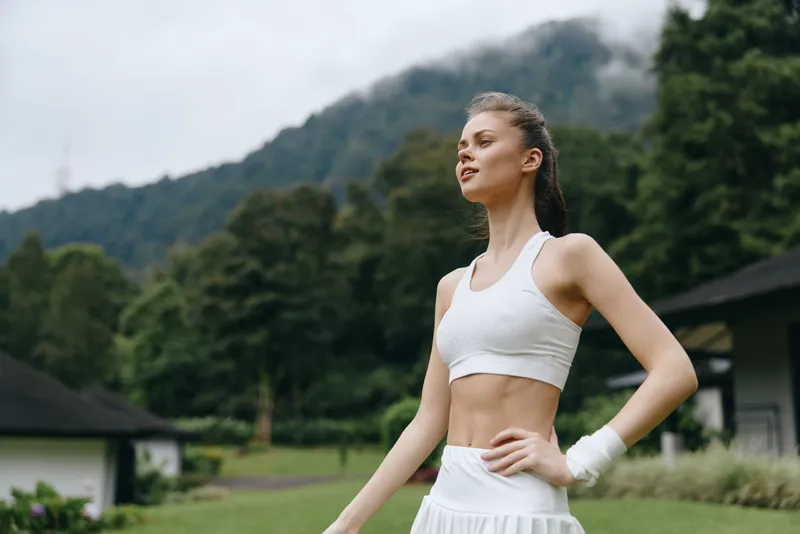 Young athletic woman in white sportswear enjoying a workout in a serene outdoor setting with lush greenery and mountains, embodying a healthy lifestyle and fitness enthusiasm