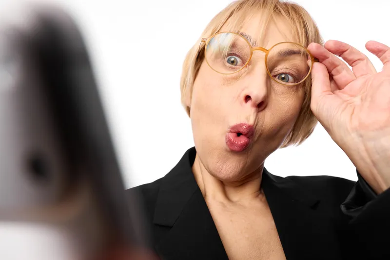Surprised senior businesswoman adjusts eyeglasses with one hand, blonde short hair, closeup face, office setting, looking at laptop screen, white background