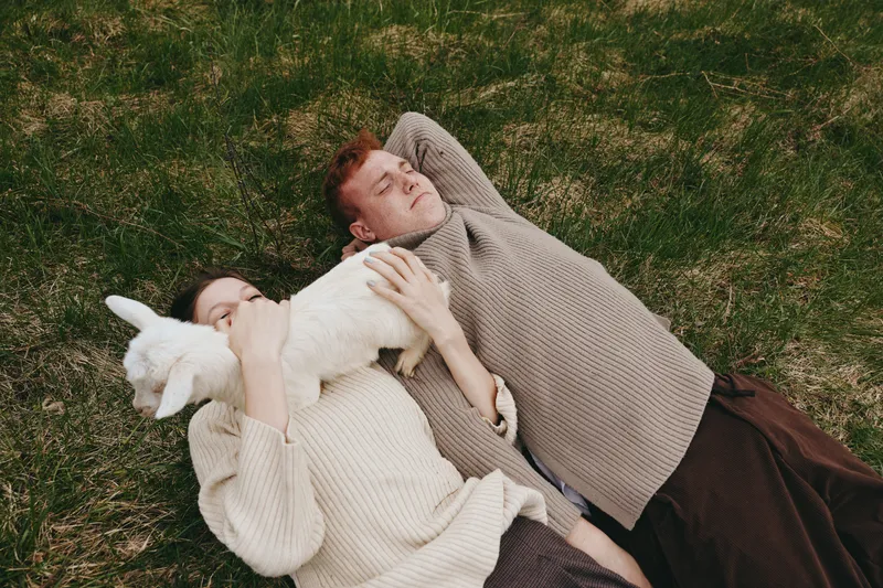 Calm couple lying on grass outdoors, cuddling a white goat kid, creating a wholesome lifestyle moment. Nature, relaxation, affection, and connection with animals in a sunny meadow setting.
