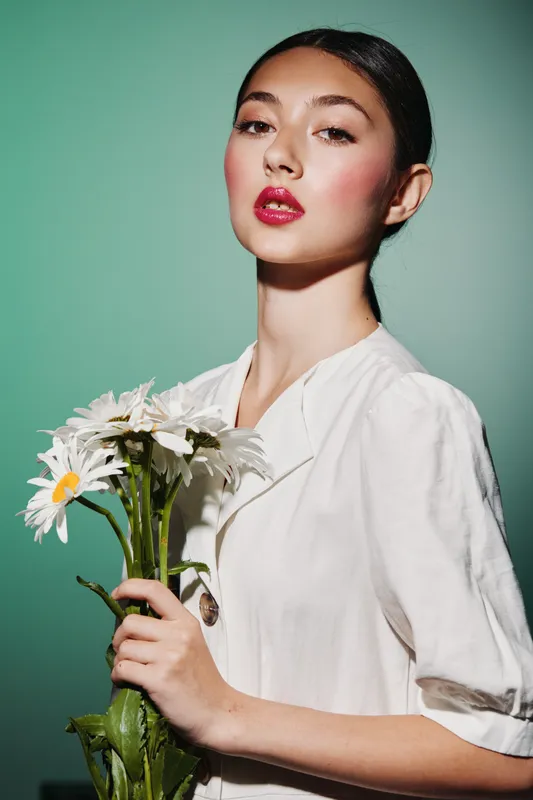 Stylish woman with vibrant makeup holds white daisies, wearing loose white clothing. Soft green background highlights beauty, fashion, and creative floral concept.