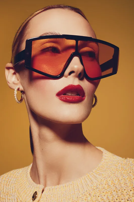 Stylish woman wearing oversized orange sunglasses and gold hoop earrings poses against warm yellow background. Fashion portrait with glossy red lips and sleek hairstyle highlights modern accessories.