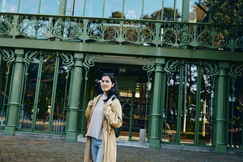 Female in a tan coat posing in front of a green glass building, urban fashion and street style, architectural facade and reflective windows, casual confidence in city setting
