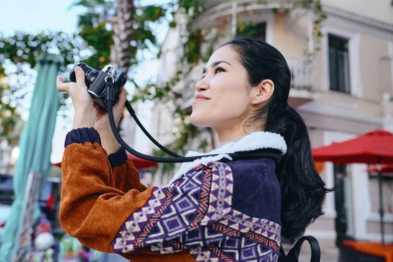Camera in hand, woman photographs a lively street scene on a sunny day, wearing a colorful knit sweater, exploring an urban environment and capturing candid moments for stock photography and creative