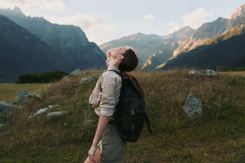 Young woman hiking in mountains, standing on grassy field, looking up with closed eyes, backpack on shoulders, enjoying nature, peaceful outdoor lifestyle moment